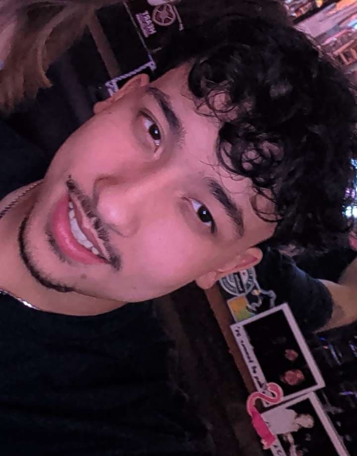 A smiling young man with dark curly hair and a goatee under pink ambient lighting.